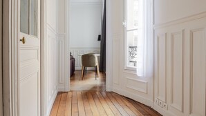 A hallway with wooden flooring, white walls, and a door leading to a room with a chair and a table.