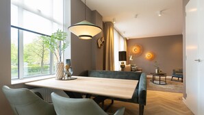 Et moderne spiseområde med et træbord, to polstrede stole og et stort vindue med udsigt til grøn bevoksning.