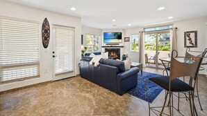 A living room with a dark blue sofa, a glass coffee table, and a fireplace. There is a TV mounted above the fireplace and a dining area with a glass table and chairs. - Beach Perfection | Private Spa, Rooftop Views, Outdoor Kitchen & Garage Parking (San Diego)
