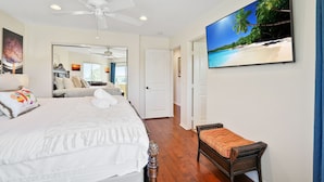 A bedroom with a bed, a bedside table, a chair, a ceiling fan, a large window, and a painting of a beach scene. - Beach Perfection | Private Spa, Rooftop Views, Outdoor Kitchen & Garage Parking (San Diego)
