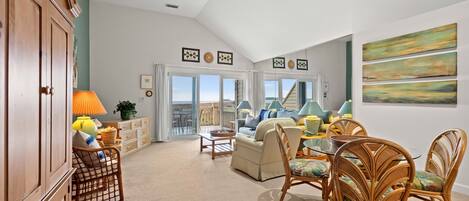 A spacious living room with a large window, a dining area with wicker chairs, and a wooden cabinet.
