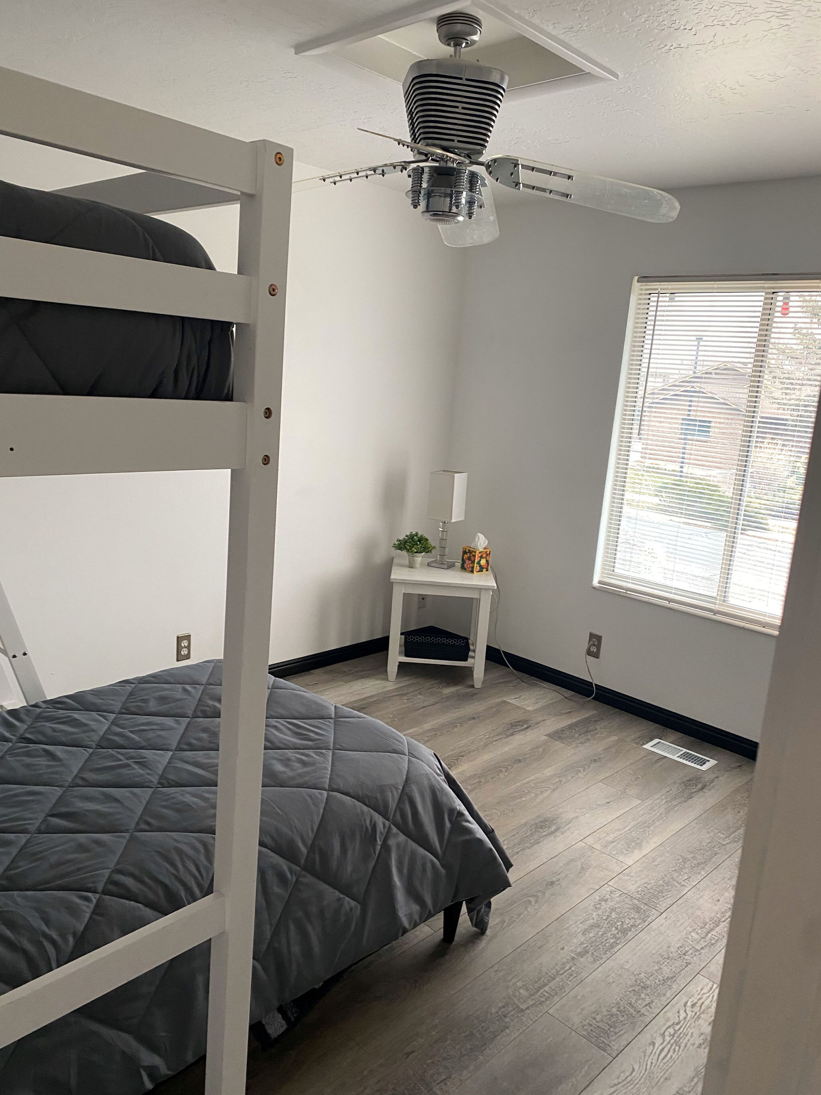 Un dormitorio con una cama doble, una mesita de noche pequeña con una lámpara, una ventana con persianas y un ventilador de techo.