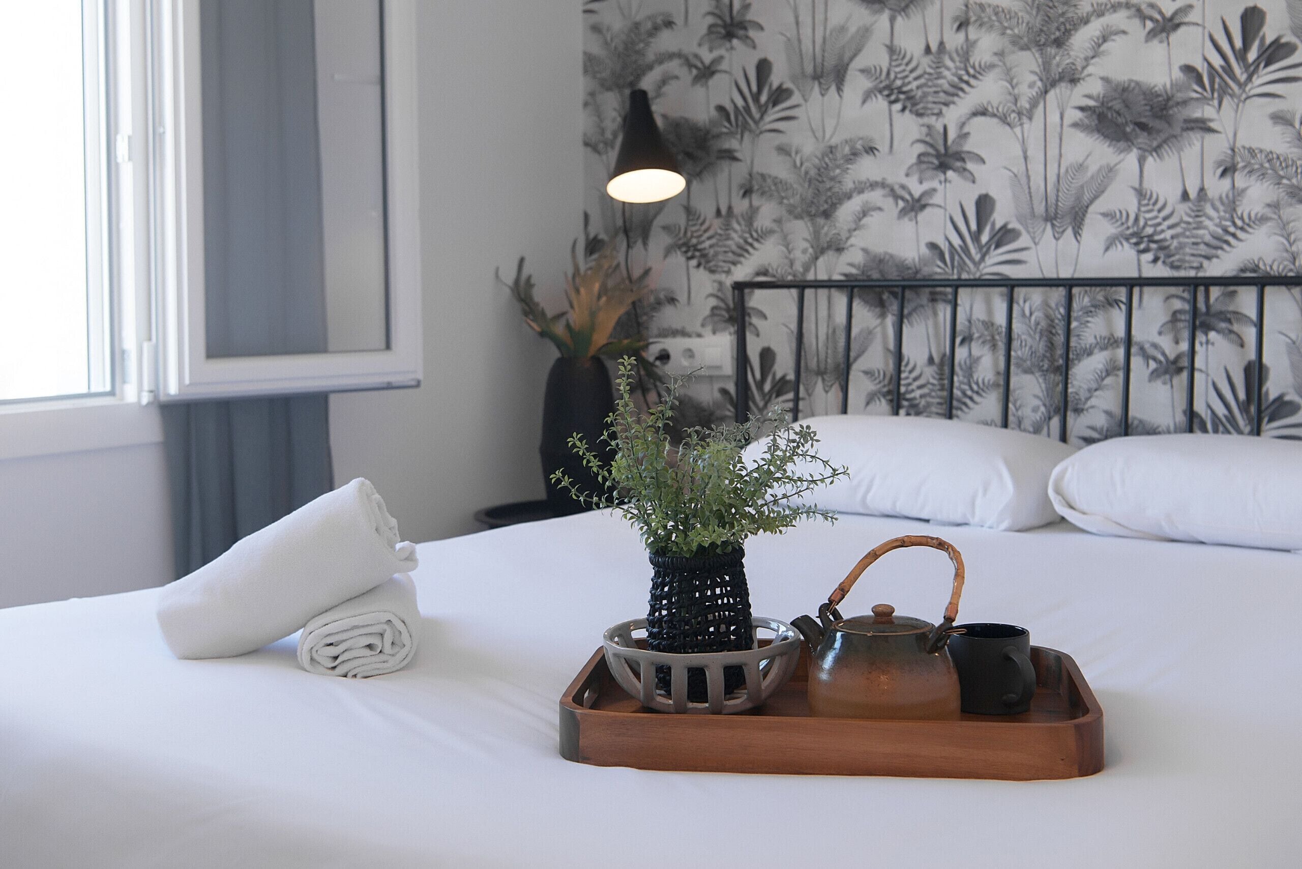Een netjes opgemaakt bed met witte lakens, een opgerolde handdoek, een dienblad met theepot en kopje, en een bloempot met plant.