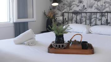 Een netjes opgemaakt bed met witte lakens, een opgerolde handdoek, een dienblad met theepot en kopje, en een bloempot met plant.