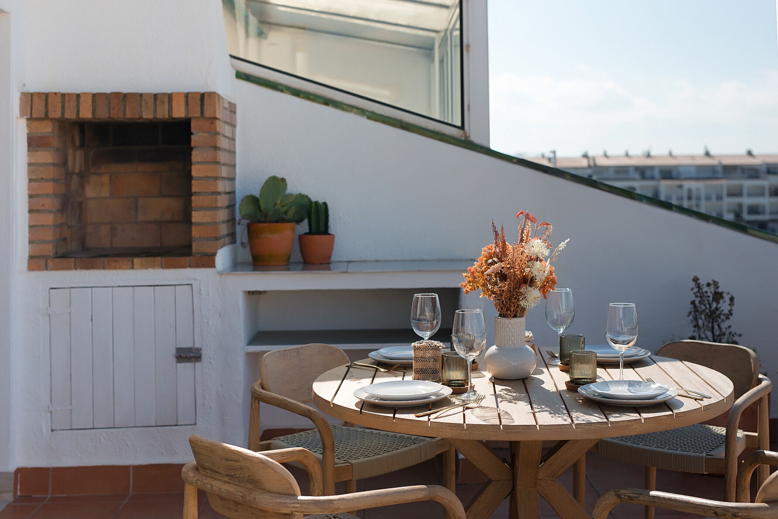 A round wooden table set for two with plates, cutlery, and glasses, accompanied by a vase of dried flowers. There are potted plants on a shelf above the table, and a brick fireplace is visible to the left.
