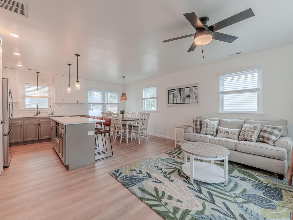 A modern kitchen with white cabinets, a dining area with a table and chairs, a sofa, and a ceiling fan.