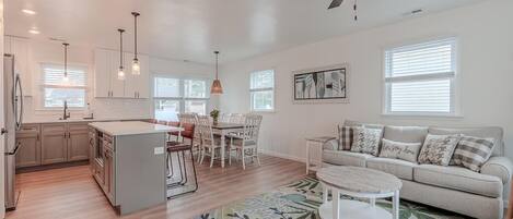 A modern kitchen with white cabinets, a dining area with a table and chairs, a sofa, and a ceiling fan.