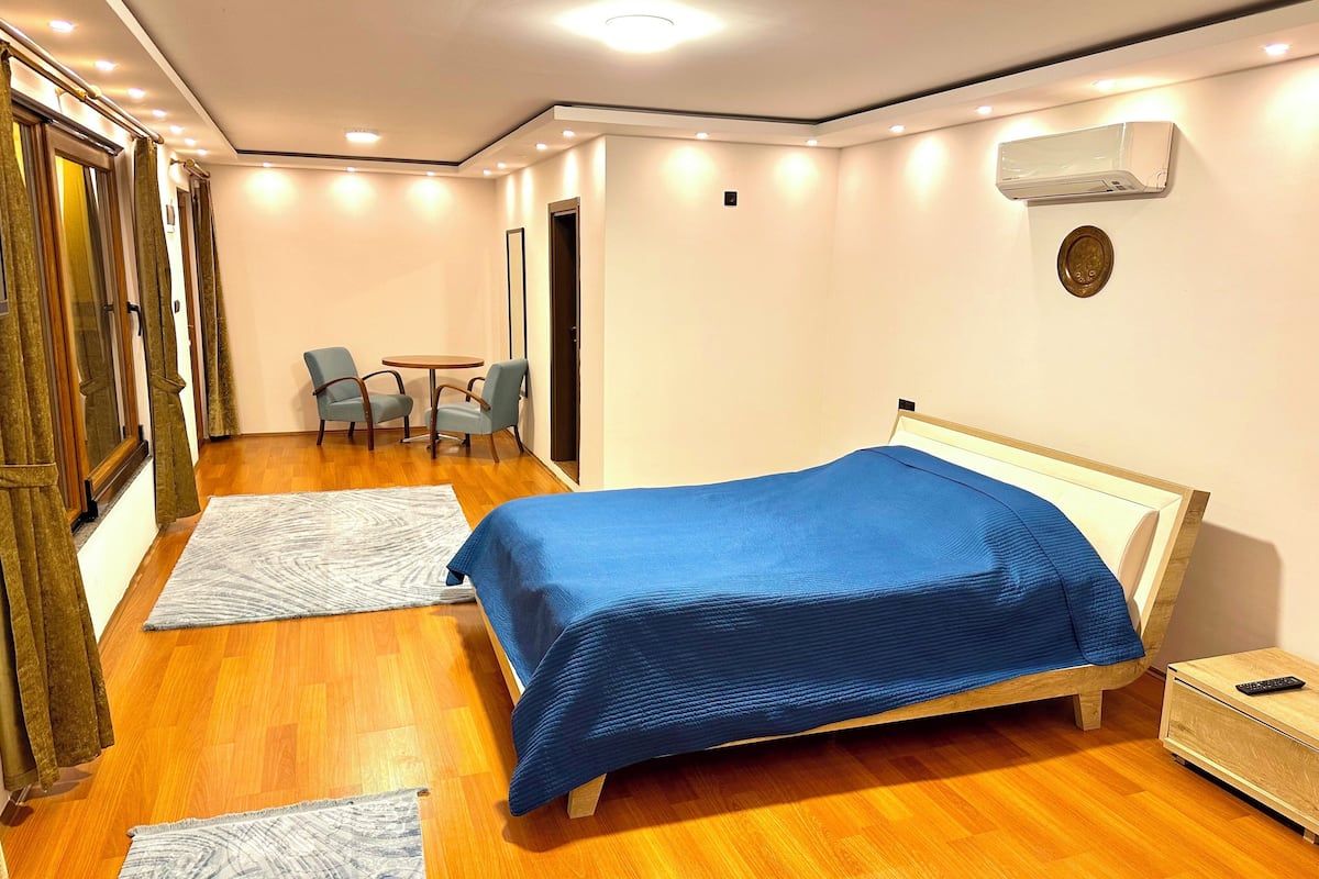 A bedroom with a bed, a bedside table, a chair, a small table, and an air conditioning unit.