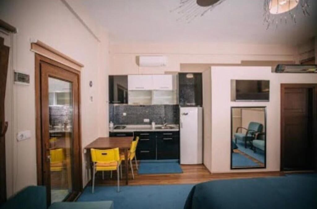 A compact room with a kitchenette, a dining area with yellow chairs, and a bed with a blue headboard.