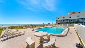 A pool area with a clear view of the ocean, a white fence, and a building with balconies.