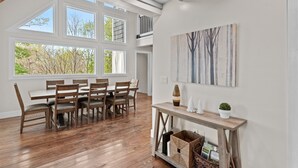 A dining area with a wooden table and chairs, large windows with a view of trees, and a wall-mounted painting. - Elevation Escape - Beech Mtn home with game room, outdoor space (Beech Mountain)