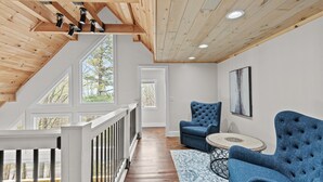 A spacious loft with a wooden ceiling, large windows, and a view of trees. There are two blue upholstered chairs and a small round table. - Elevation Escape - Beech Mtn home with game room, outdoor space (Beech Mountain)