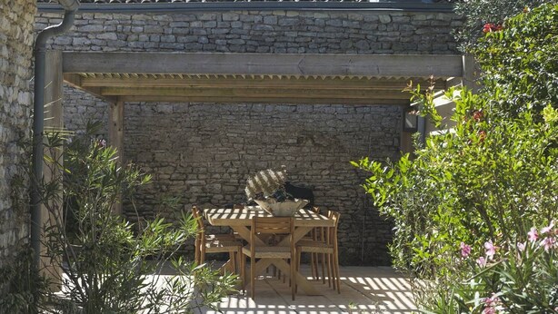 Un edificio de piedra con tejado de baldosas, una pérgola de madera y una mesa con sillas debajo.