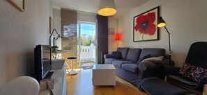 Un salon moderne avec un canapé gris, une petite table blanche, un lampadaire et une grande fenêtre donnant sur un balcon.