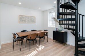 Un espace repas comprenant une table et des chaises en bois, une console noire, un escalier en colimaçon et une œuvre d’art encadrée accrochée au mur.