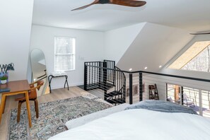 Une chambre avec un grand lit, un bureau avec une chaise, une petite table avec une plante, un miroir, une fenêtre avec des stores, un escalier avec une rampe et un ventilateur de plafond.
