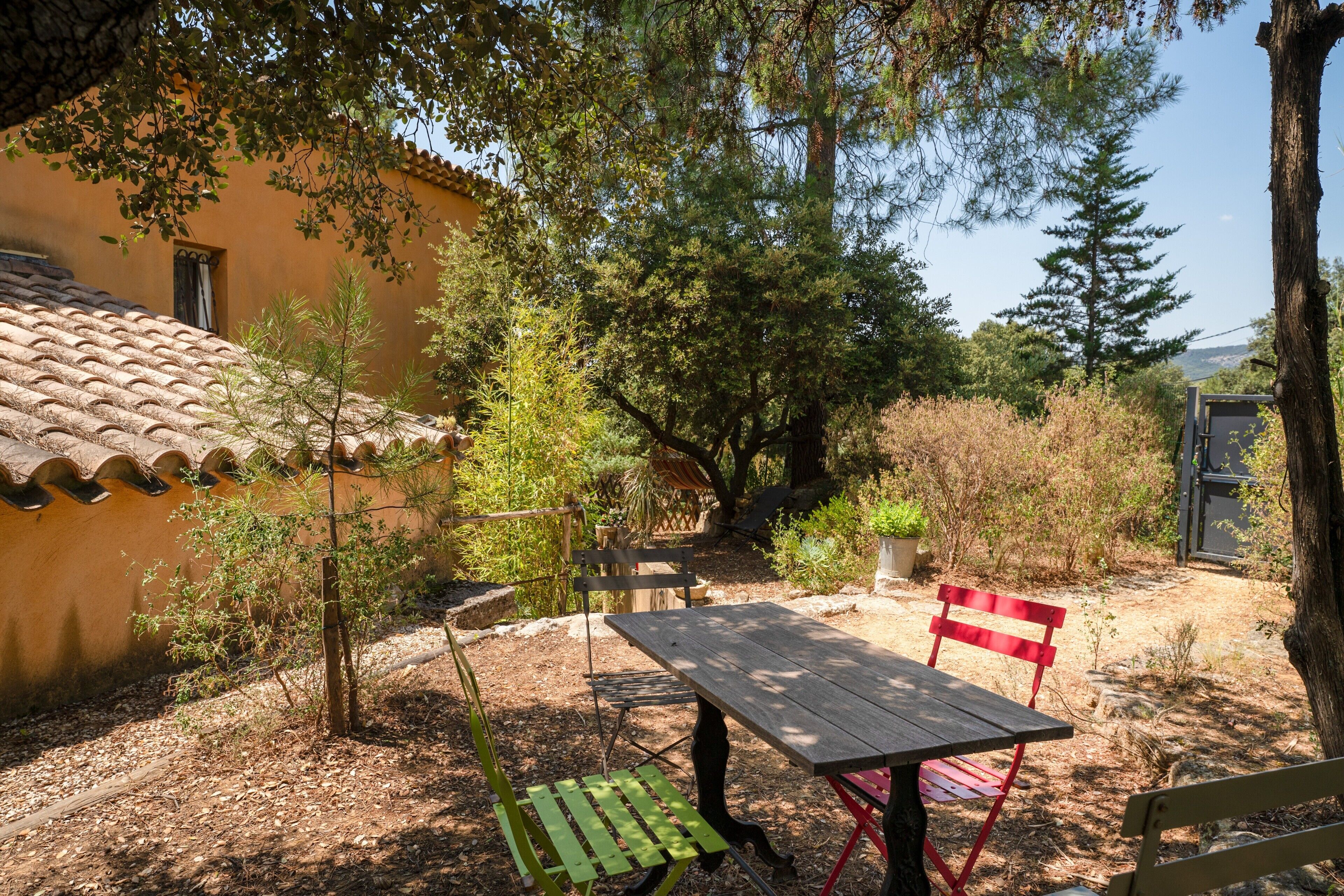 An outdoor patio with a wooden table and chairs, surrounded by trees and a building with a tiled roof.