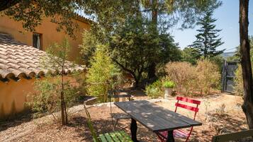 An outdoor patio with a wooden table and chairs, surrounded by trees and a building with a tiled roof.