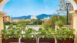A terrace with wicker chairs and tables, potted plants, and a view of buildings and mountains. - Apartment 'Mazzini' with Sea View, Wi-Fi, and Air Conditioning (Golfo Aranci)