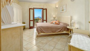 A bedroom with a bed, a bedside table, a lamp, a chair, a dresser, and a view of the outside. - Apartment 'Mazzini' with Sea View, Wi-Fi, and Air Conditioning (Golfo Aranci)