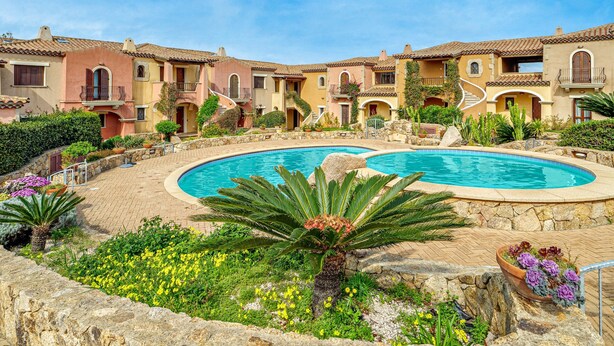A pool surrounded by multi-story buildings with balconies, a stone wall, and various plants. - Apartment 'Mazzini' with Sea View, Wi-Fi, and Air Conditioning (Golfo Aranci)