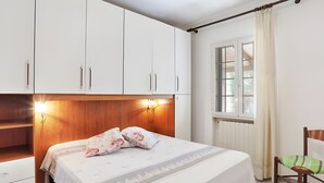 A bedroom with a bed, a wooden headboard, white cabinets, a chair, and a window with curtains. - Apartment 'Tramonto' with Private Terrace, Wi-Fi and Air Conditioning (Capoliveri)