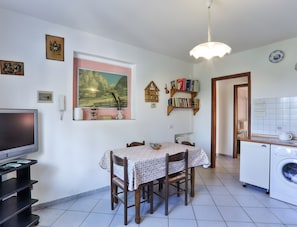 A dining area with a table, chairs, a television, and a kitchen with a washing machine.