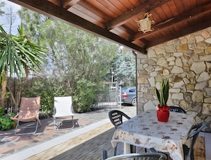 Une terrasse couverte avec un mur en pierre, une table recouverte d’une nappe à motifs floraux et des chaises.