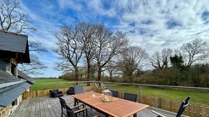Una terraza de madera con una mesa y sillas, vista a un campo verde y árboles sin hojas.