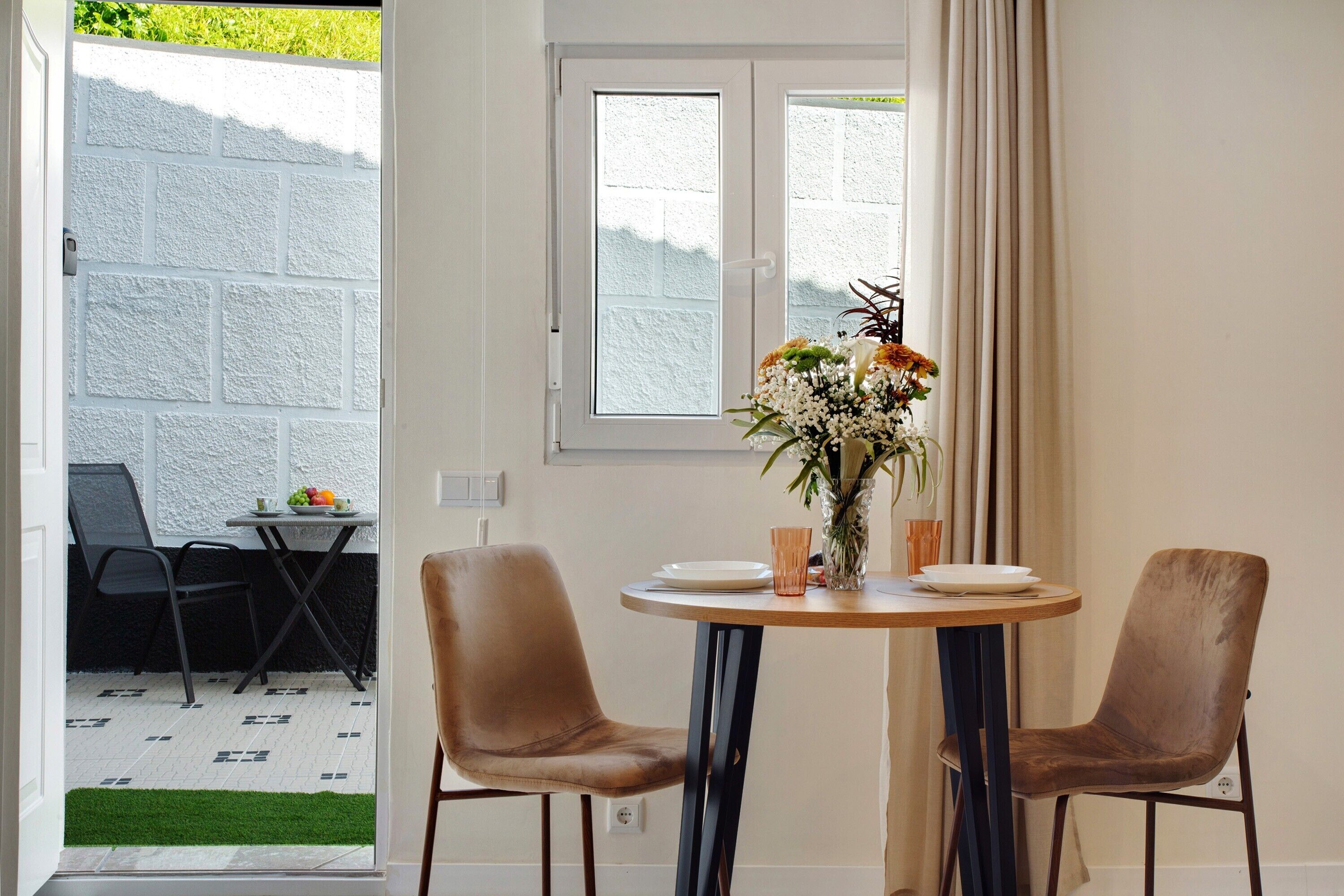 Un comedor con una mesa redonda, dos sillas, un florero con flores y una ventana con cortinas.