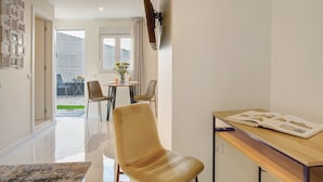 A modern living room with a sofa, a dining table with chairs, a TV mounted on the wall, and a view of the outdoors.