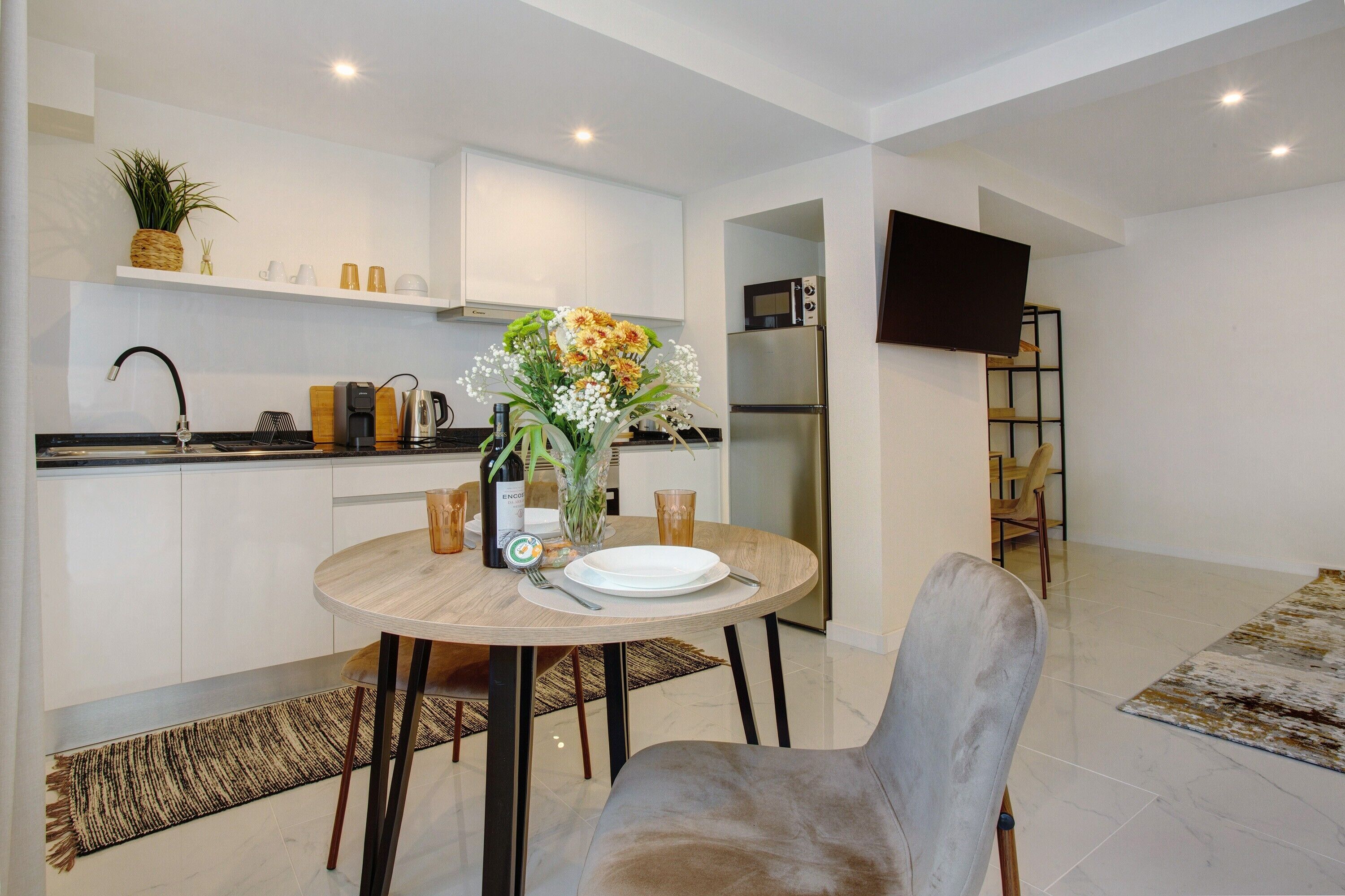 A modern kitchen with white cabinets, a dining table with a vase of flowers, and a flat-screen TV mounted on the wall.