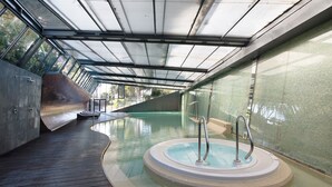 Een modern binnenzwembad met glazen wanden, een houten terras en een jacuzzi.