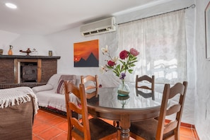 Un coin repas comprenant une table ronde en verre, des chaises en bois, une cheminée, un canapé et un tableau représentant un coucher de soleil.