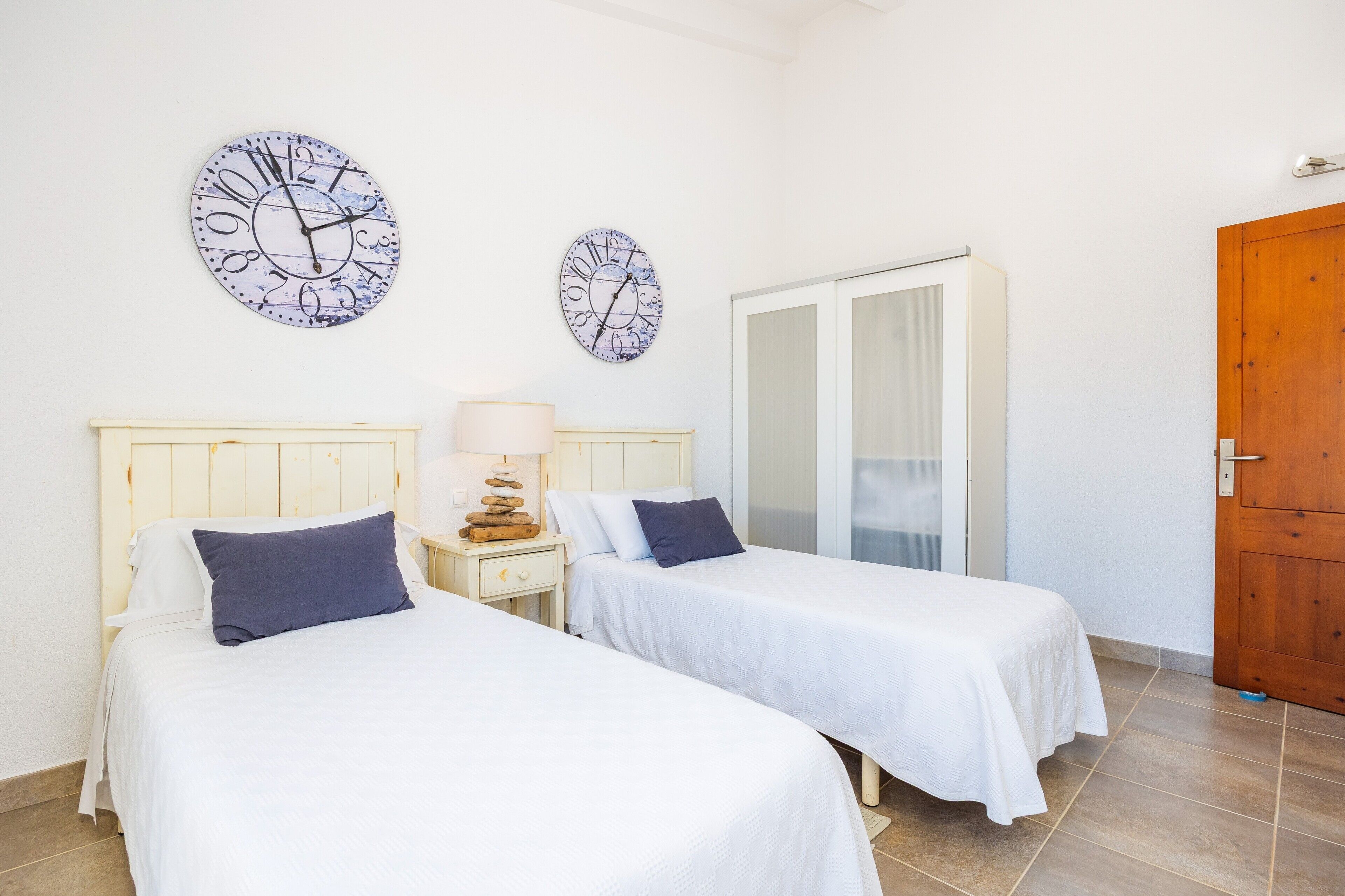 A room with two beds, a wooden wardrobe, and two wall clocks.
