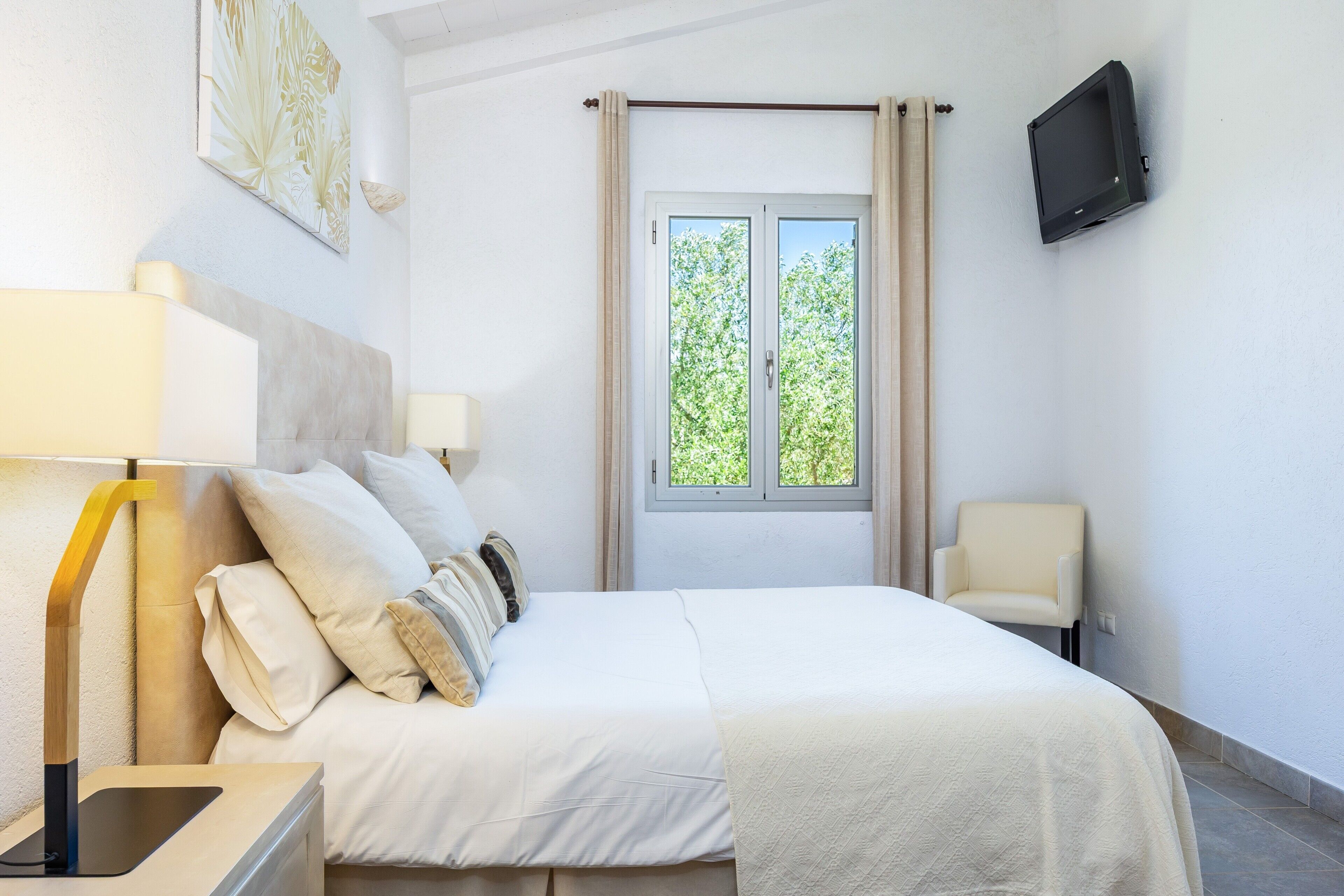 Una habitación de hotel con cama, sillón, televisor, ventana con cortinas y lámpara.