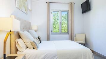 Una habitación de hotel con cama, sillón, televisor, ventana con cortinas y lámpara.