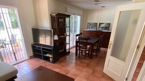 Una sala de estar con televisor, una mesa de centro de madera, una mesa de comedor y sillas. Hay una puerta corrediza de vidrio que da a un patio.