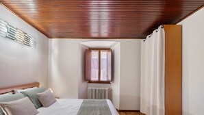 Un dormitori amb sostre de fusta, una finestra amb cortines, un radiador i un llit amb almoades.