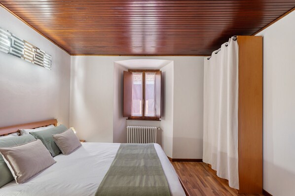 A bedroom with a wooden ceiling, a window with curtains, a radiator, and a bed with pillows.