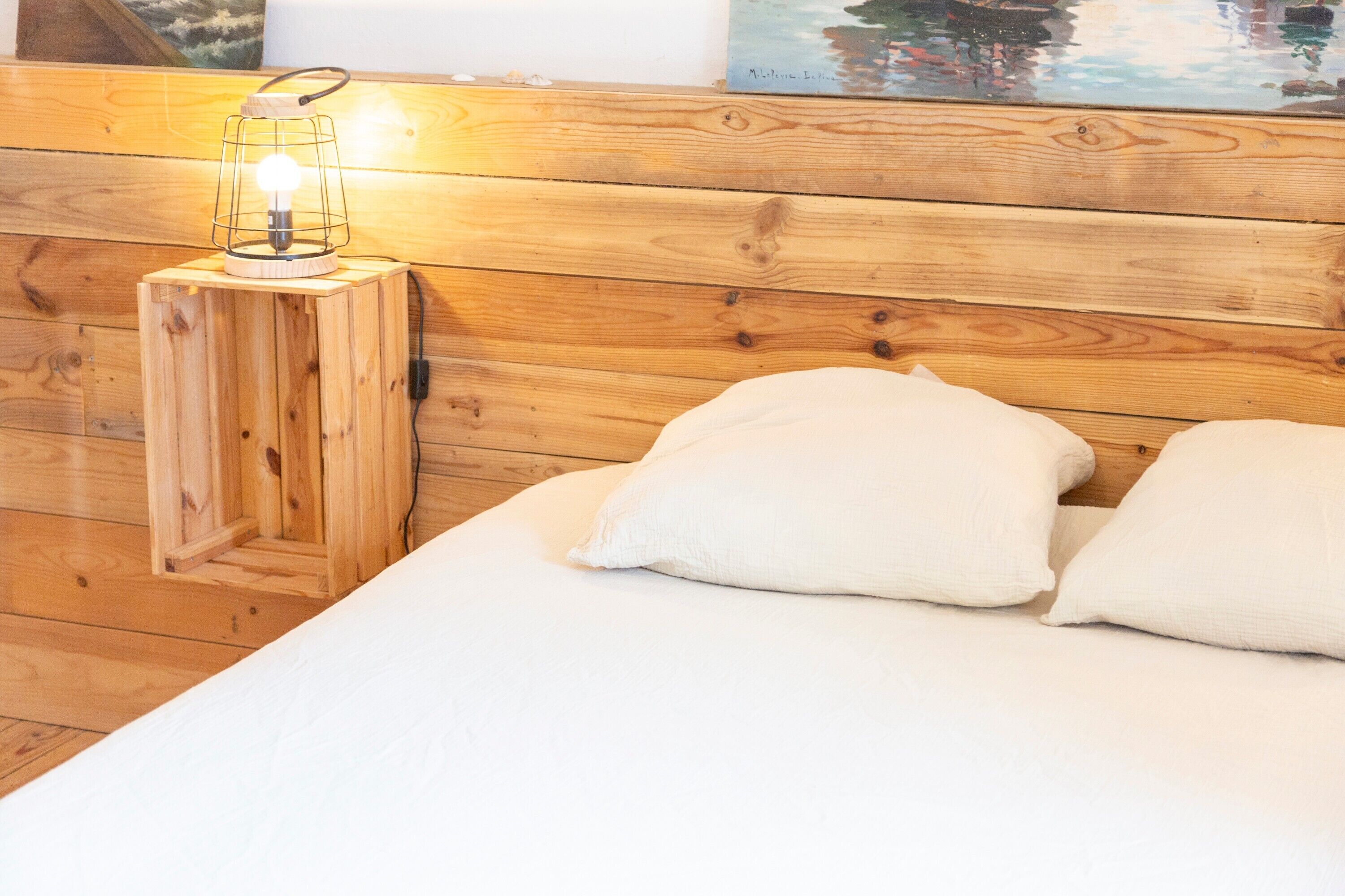 A wooden headboard with a small wooden stool holding a light, a white bed with pillows, and a painting above the bed.