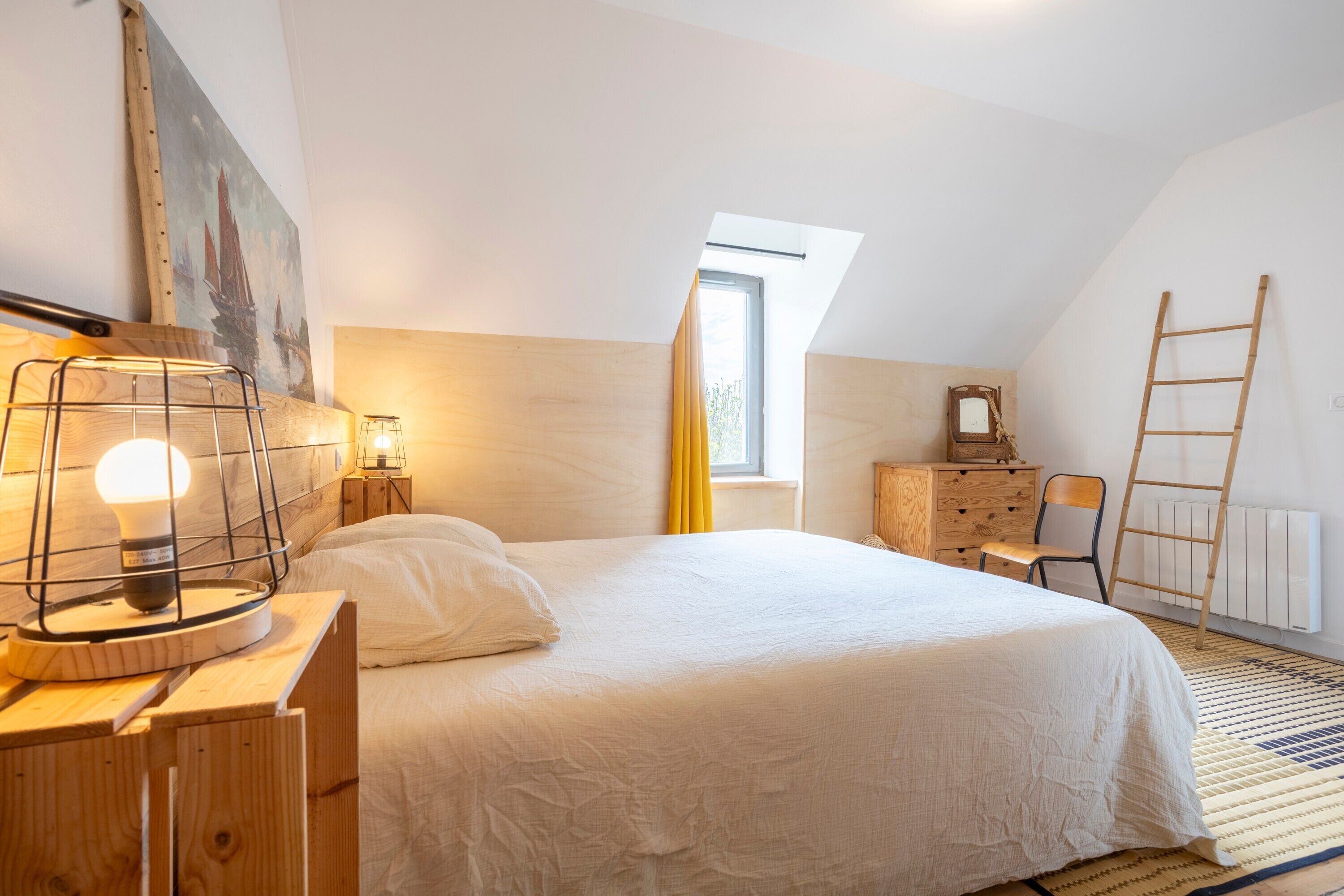 A bedroom with a bed, a wooden headboard, a nightstand with a lamp, a dresser, a chair, a ladder, a window, and a painting on the wall.