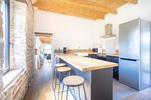 A modern kitchen with a large island, bar stools, a stainless steel refrigerator, and a stone wall.