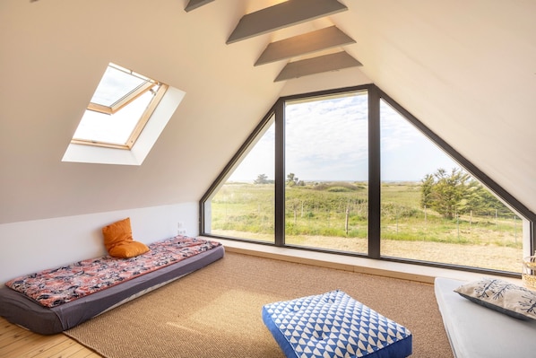 A room with a sloped ceiling, a large window with a view of a field, a sofa with patterned cushions, and a small table with a cushion.