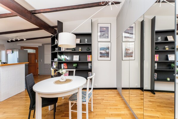 Una cocina moderna con vigas de madera, una mesa de comedor blanca y sillas negras. Hay estantes negros con libros y objetos decorativos.