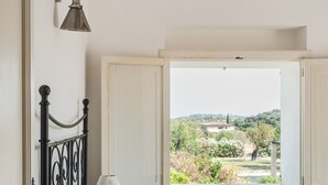 Et soveværelse med seng, natbord, lampe og et vindue med udsigt over grønne omgivelser.