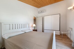 A bedroom with a bed, nightstand, lamp, chair, wardrobe, and an air conditioning unit.