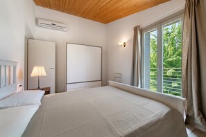 Une chambre avec un lit, une table de chevet, un appareil de climatisation, une fenêtre avec des rideaux et une vue sur la verdure à l’extérieur.