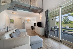 Un salon moderne avec un canapé, un pouf et un téléviseur. Il y a également un coin cuisine avec une table à manger et des chaises. La pièce est dotée de grandes fenêtres et d’un balcon avec des sièges d’extérieur.