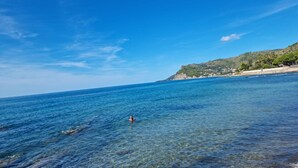 A coastal scene with clear blue water, a person swimming, and a hilly landscape with buildings in the background. - Villa 'With Pool in Private Park' with Sea View, Wi-Fi and Air Conditioning (Agnone Cilento)